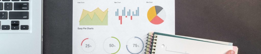 A person holds a notebook and pen next to a laptop and a printed sheet showing various colorful charts and graphs, including a line chart, bar chart, pie chart, and progress circles.